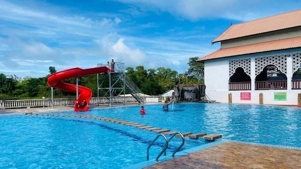A swimming pool with a red slide, a covered area with a tiled roof, and a building with a white facade and decorative arches.