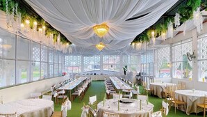 Een feestzaal met lange en ronde tafels, bedekt met witte doek en versierd met groene planten, en aan één kant grote ramen.