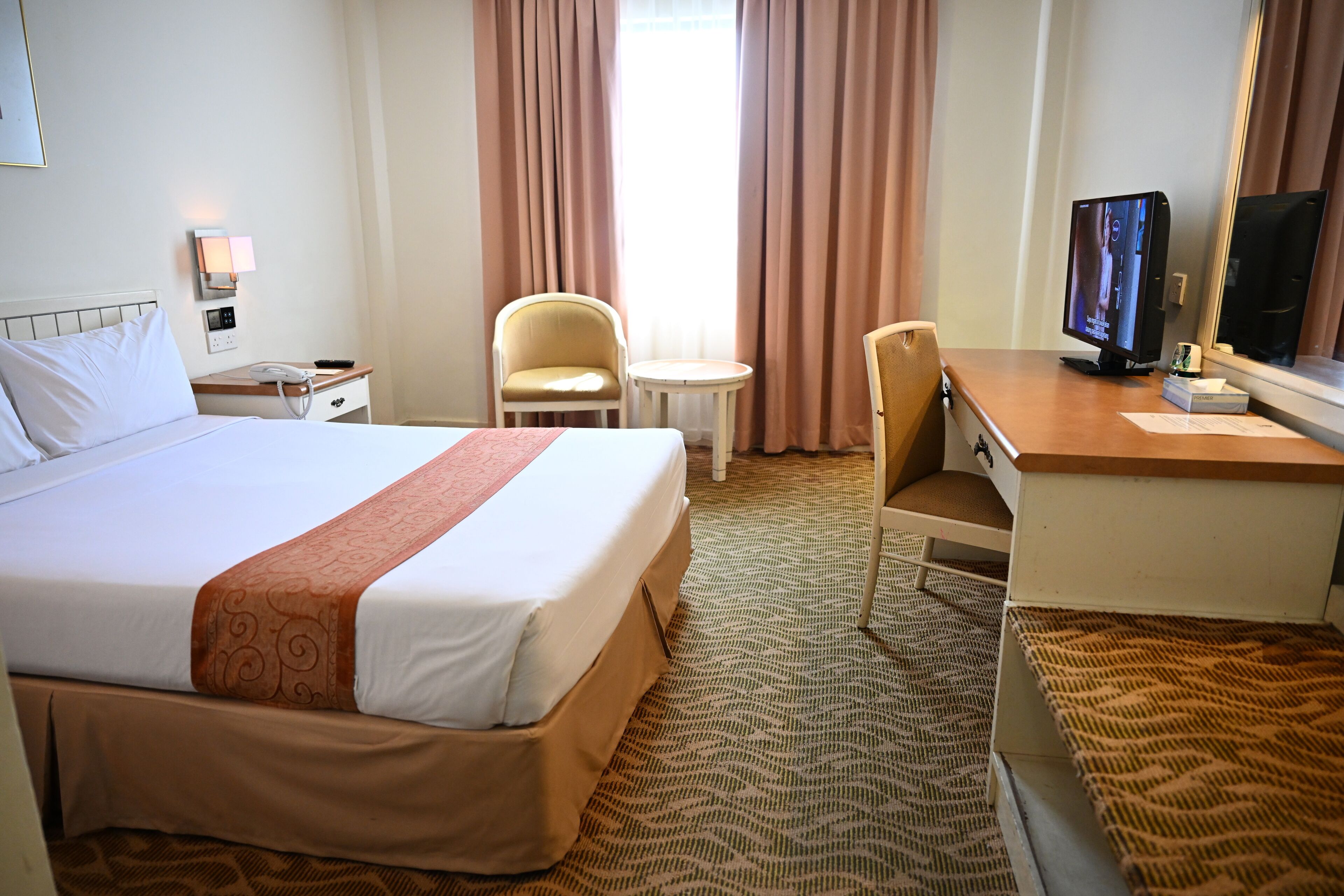 A hotel room with a large bed, a desk with a computer, a chair, a small table, and a television.