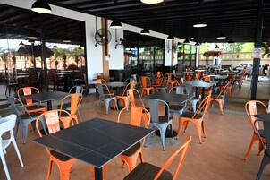 Un área de comedor al aire libre con mesas y sillas, una sección cubierta con pared de vidrio y vistas a un edificio y árboles.