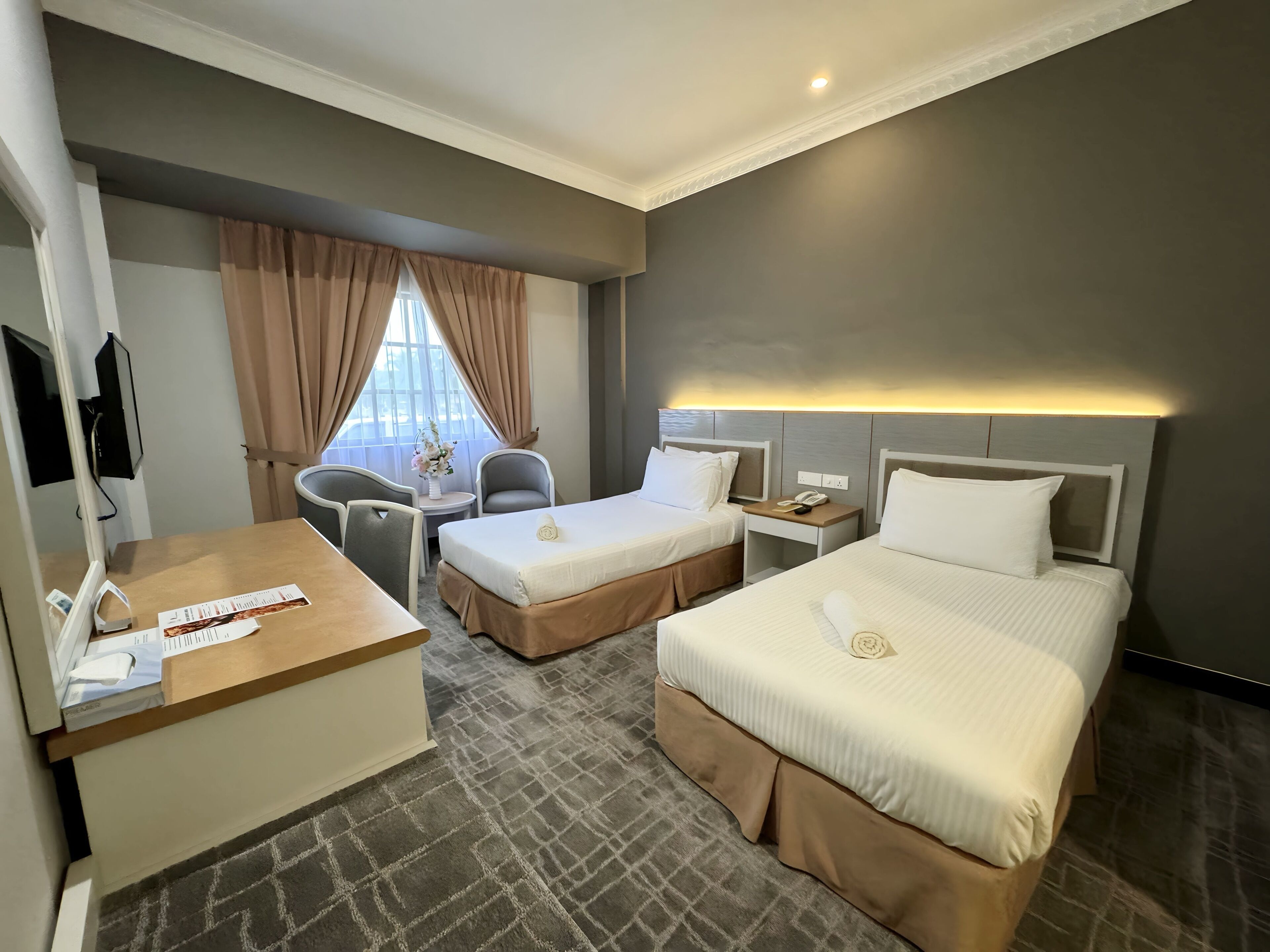 Habitación de hotel con dos camas, un escritorio, una silla, una mesita con un florero y un televisor montado en la pared.