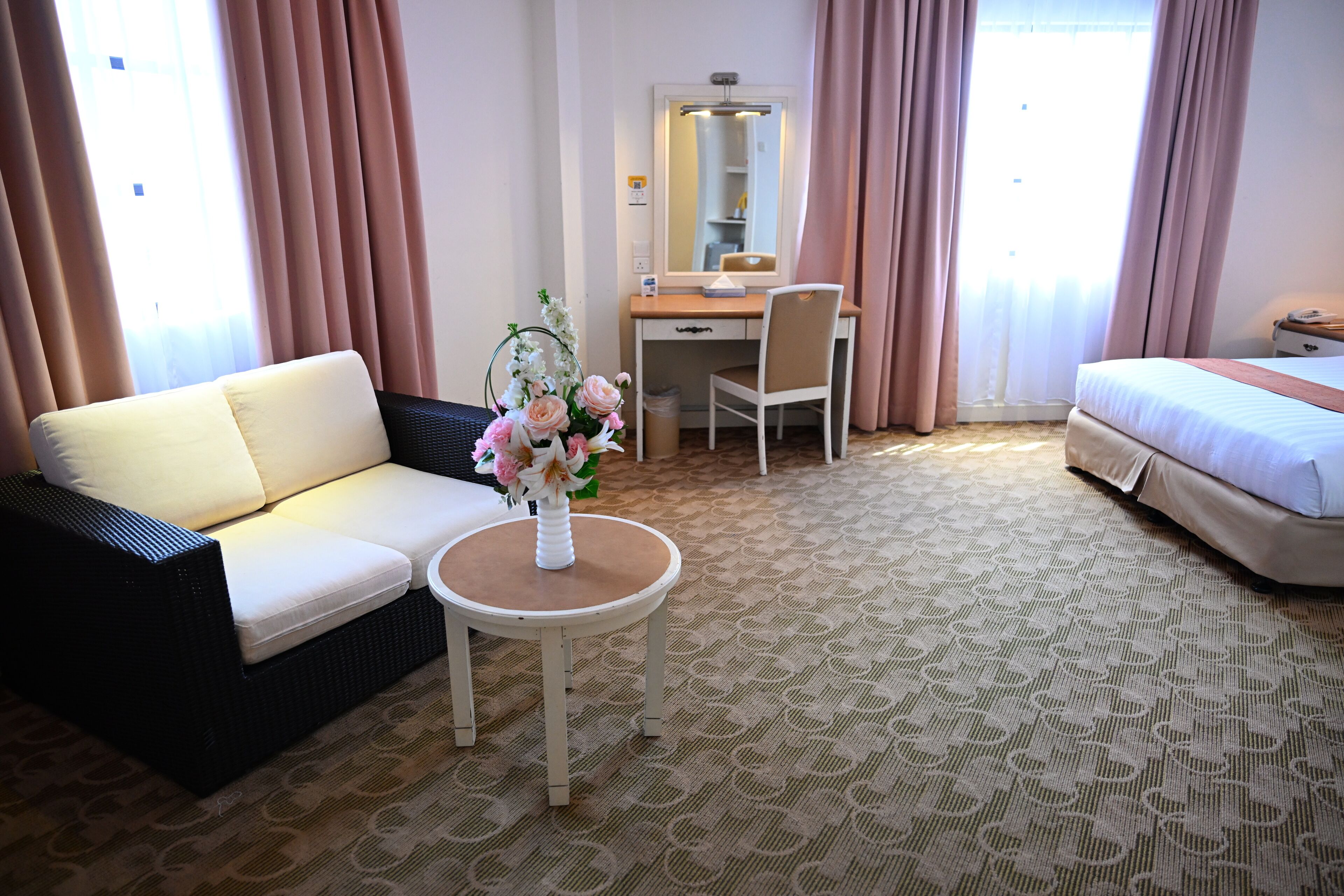 A hotel room with a sofa, a desk with a chair, a small table with a vase of flowers, a bed, and a large window with curtains.