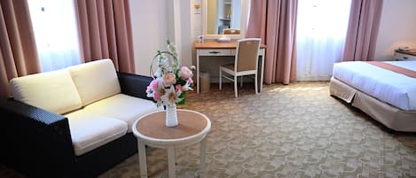A hotel room with a sofa, a desk with a chair, a small table with a vase of flowers, a bed, and a large window with curtains.