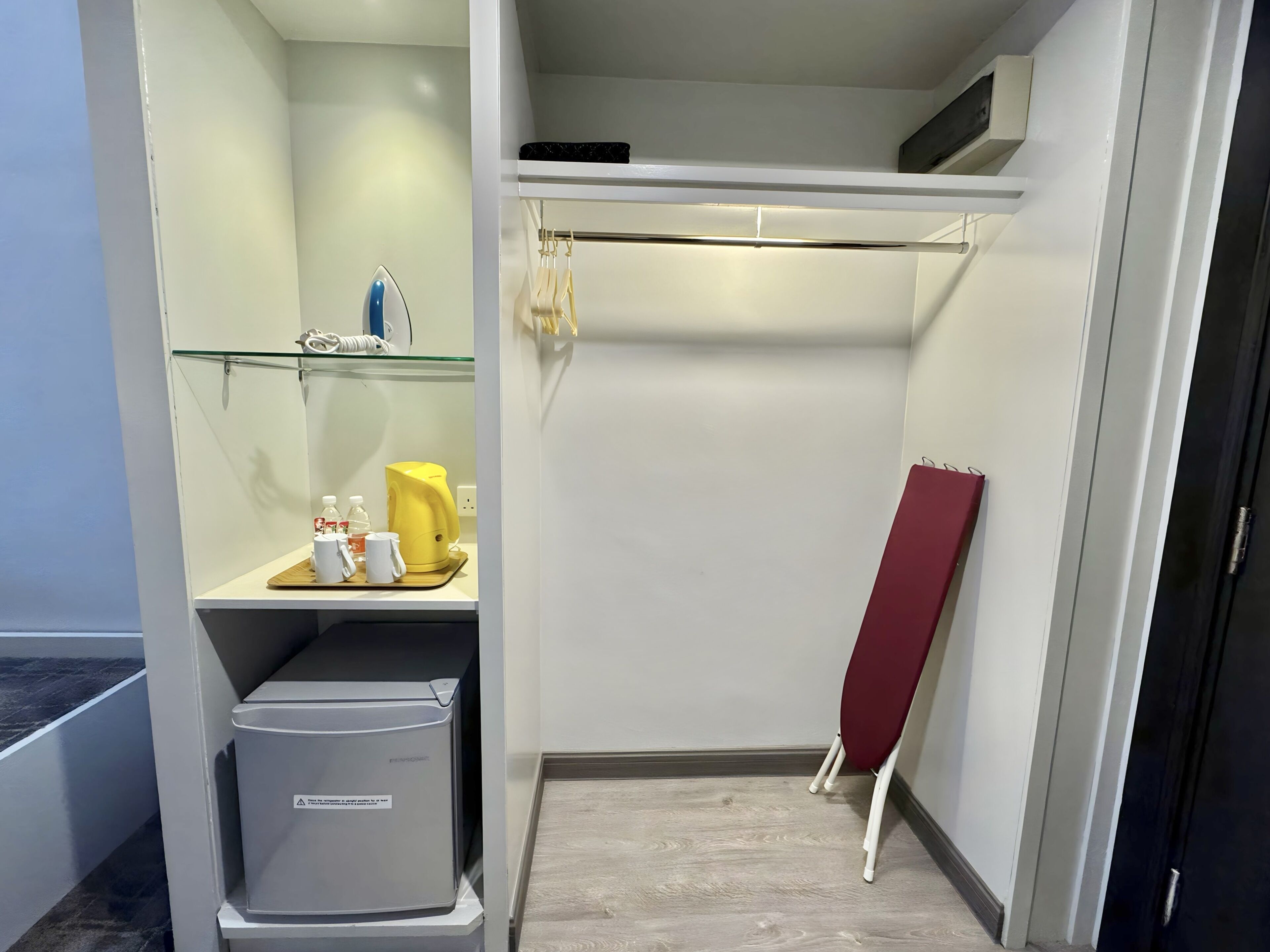 A compact hotel room with a built-in ironing station, a small shelf with toiletries, and a red folding chair.