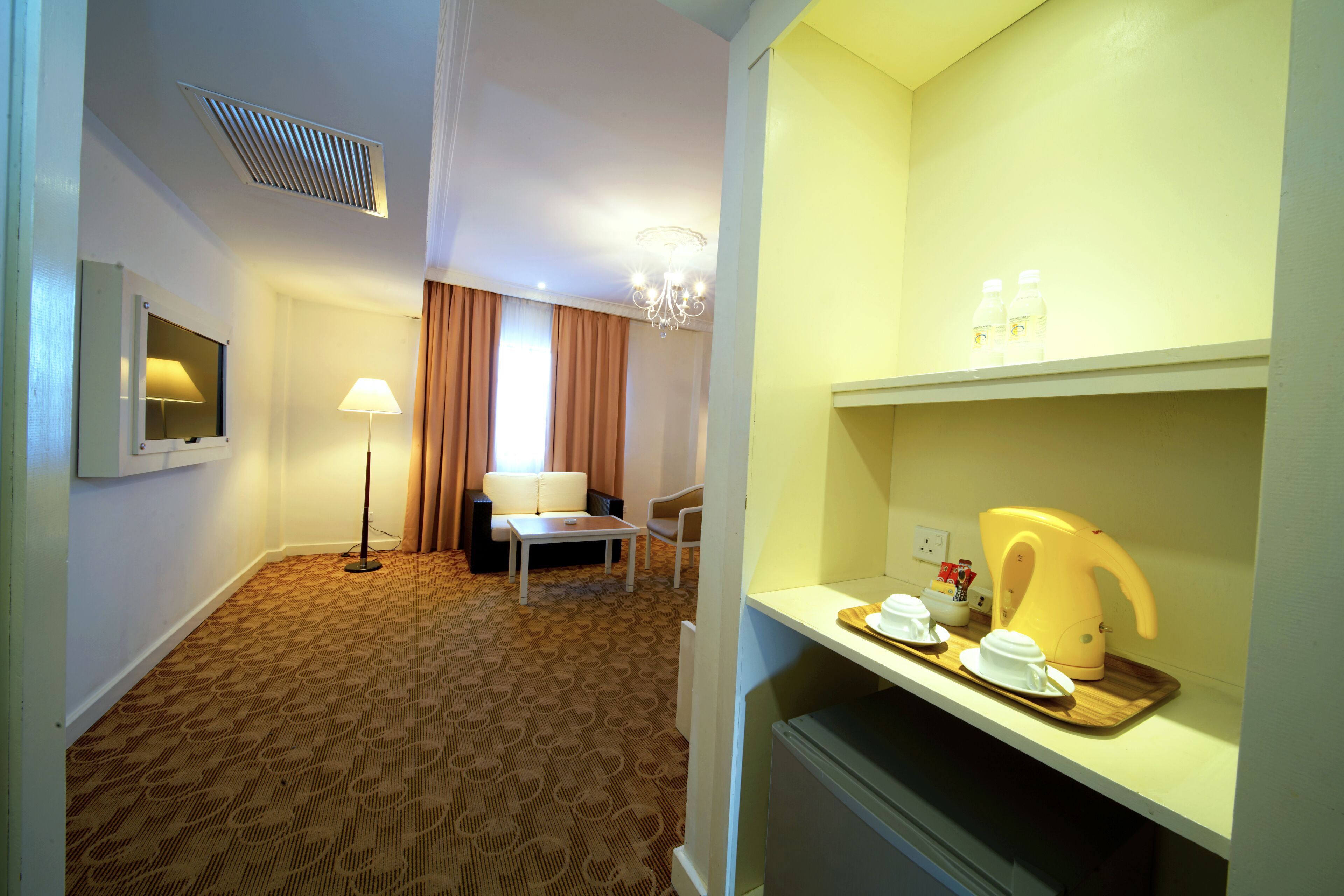 A hotel room with a patterned carpet, a sofa, a table, a lamp, a chandelier, and a breakfast bar with cups and a kettle.