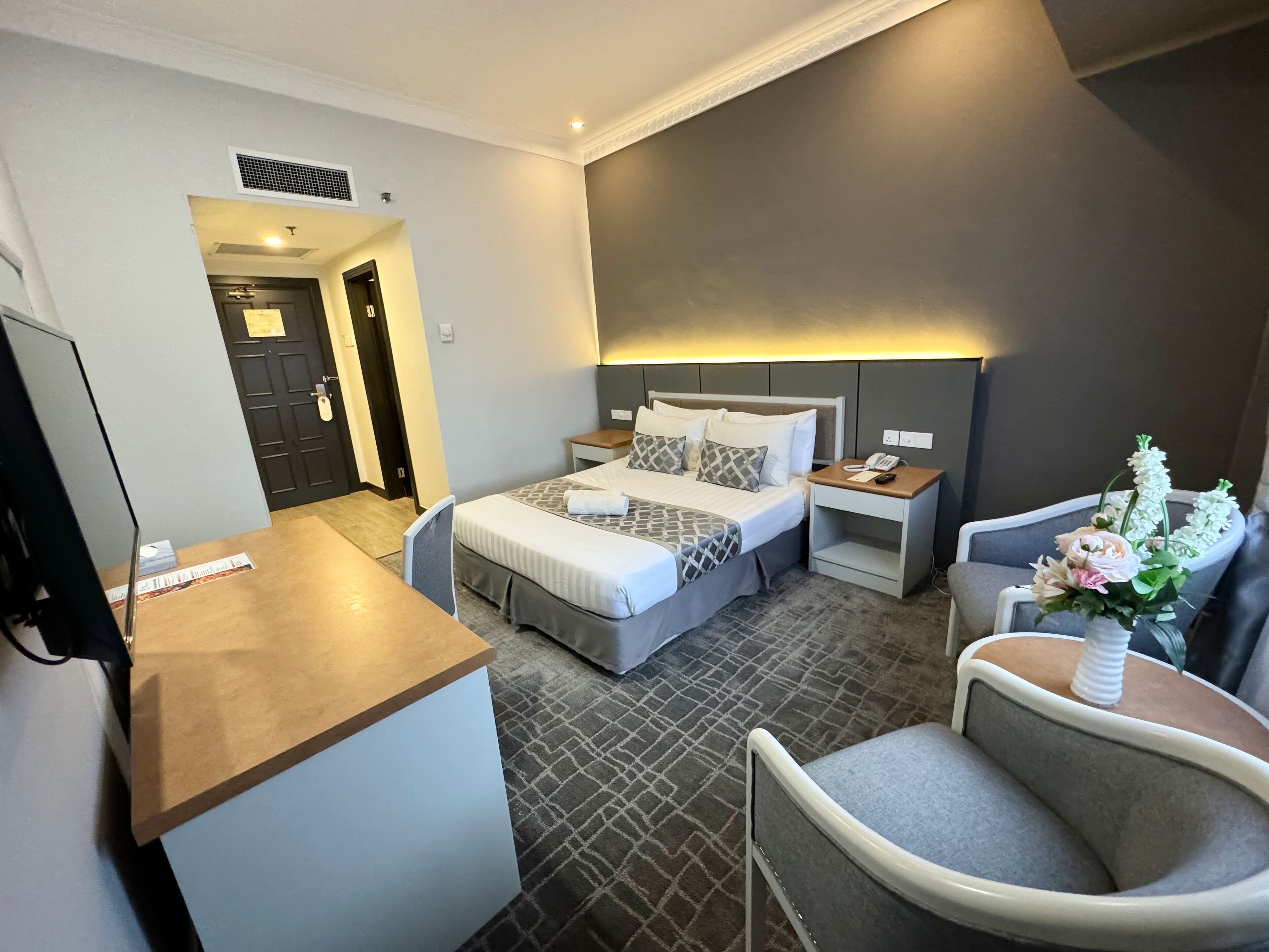 Quarto de hotel com cama, mesas de cabeceira, escrivaninha, cadeira e uma pequena mesa com um vaso de flores.
