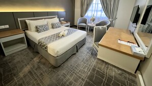 Habitación de hotel con una cama grande, mesitas de noche, un escritorio con silla, una pequeña mesa con un jarrón de flores y un televisor.