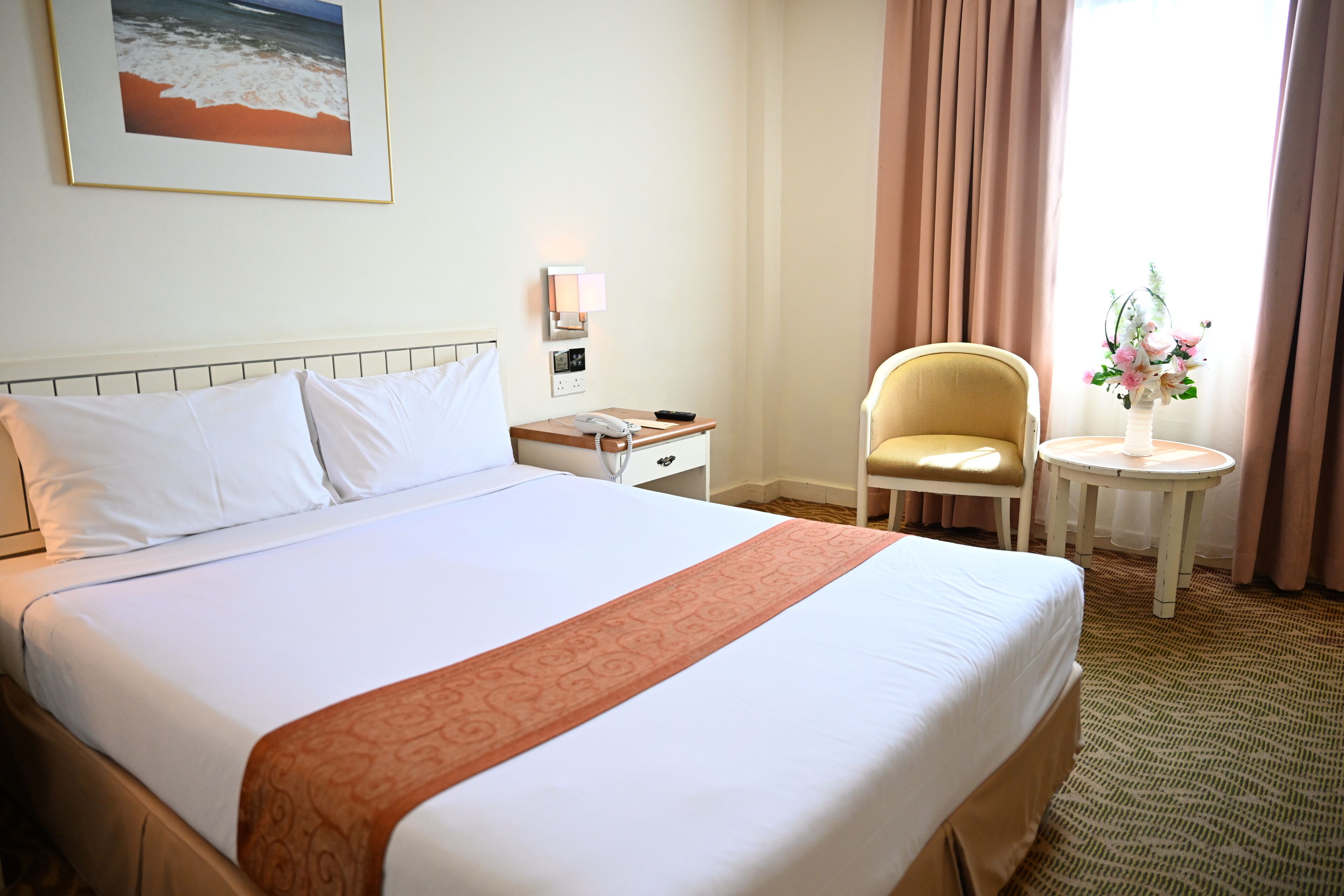A hotel room with a large bed, a bedside table with a phone, a chair, a small table with a vase, and a painting on the wall.