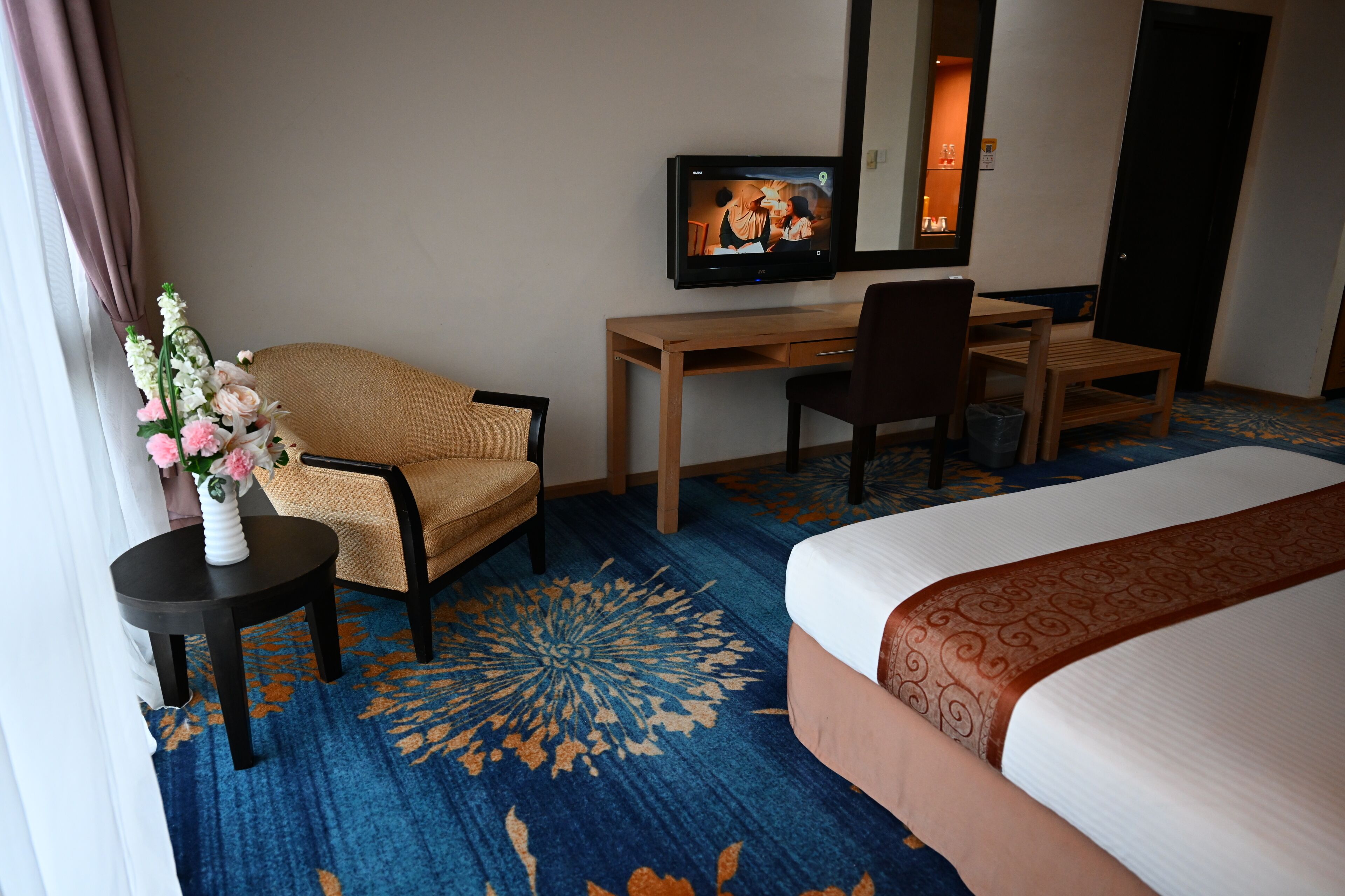 Habitación de hotel con cama, escritorio, silla, televisor y un jarrón con flores.