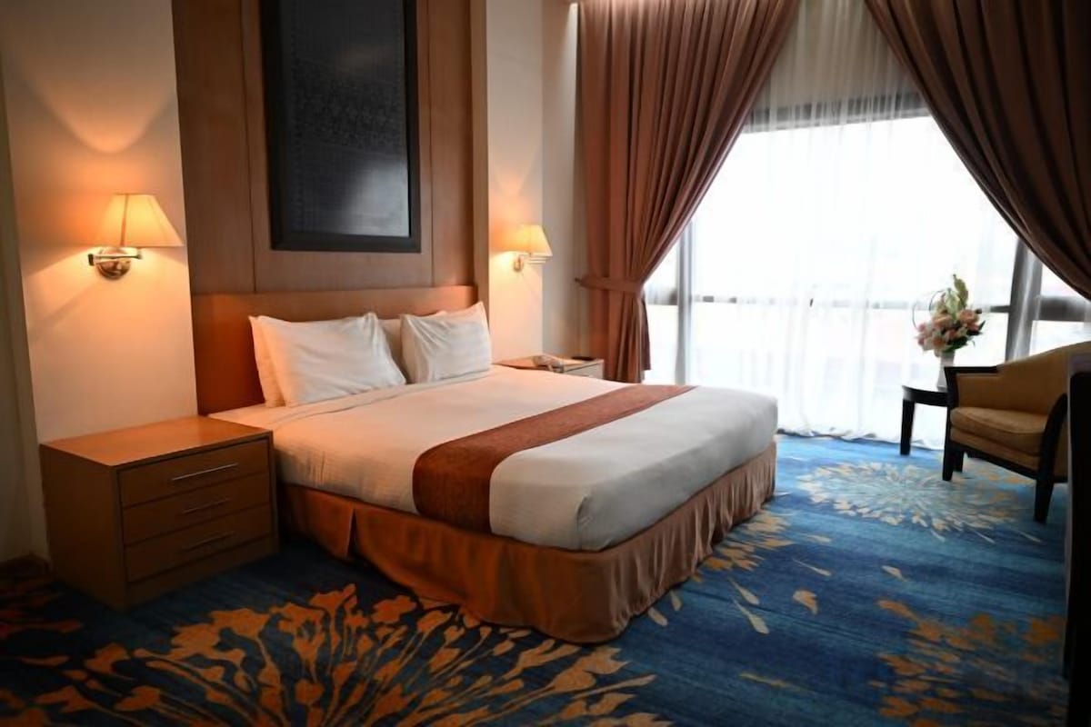 A hotel room with a large bed, two bedside tables, a nightstand, a chair, and a window with curtains.