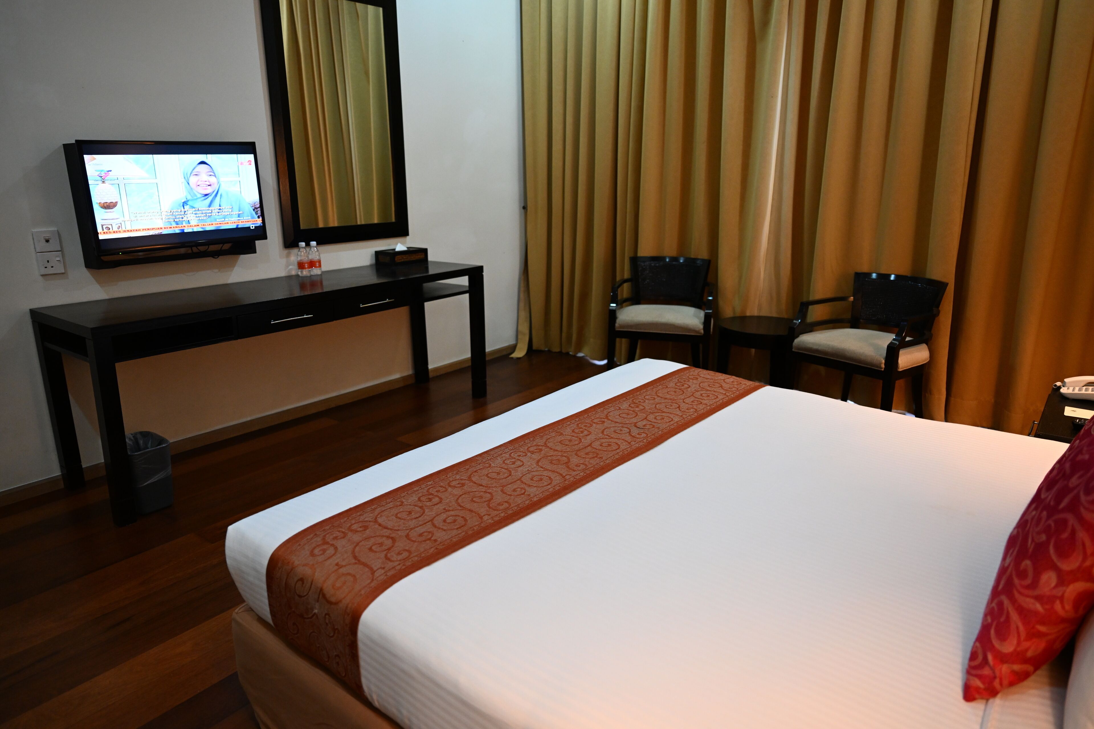 A hotel room with a bed, a desk with a television, two chairs, and a window with curtains.