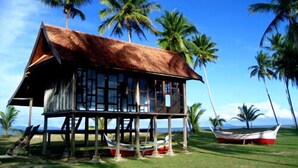 A wooden house with a thatched roof, situated on a grassy area with palm trees in the background. - Terrapuri Heritage Village (Setiu)