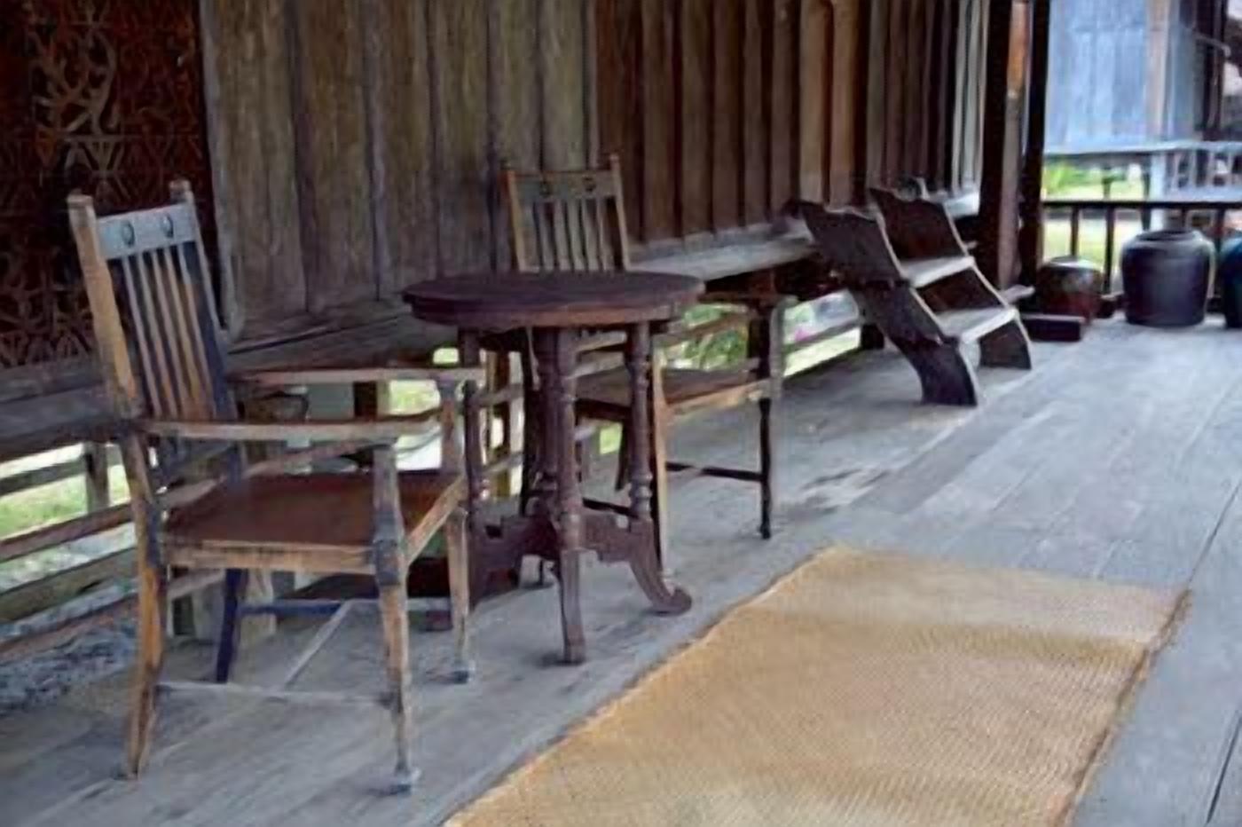 A wooden porch with rustic furniture, including a bench and chairs, and a woven mat on the floor.