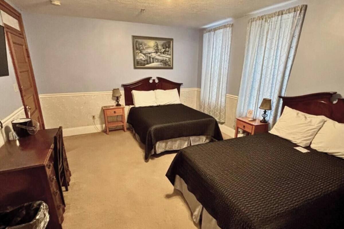 A hotel room with two beds, a wooden dresser, a painting on the wall, and a ceiling light fixture.