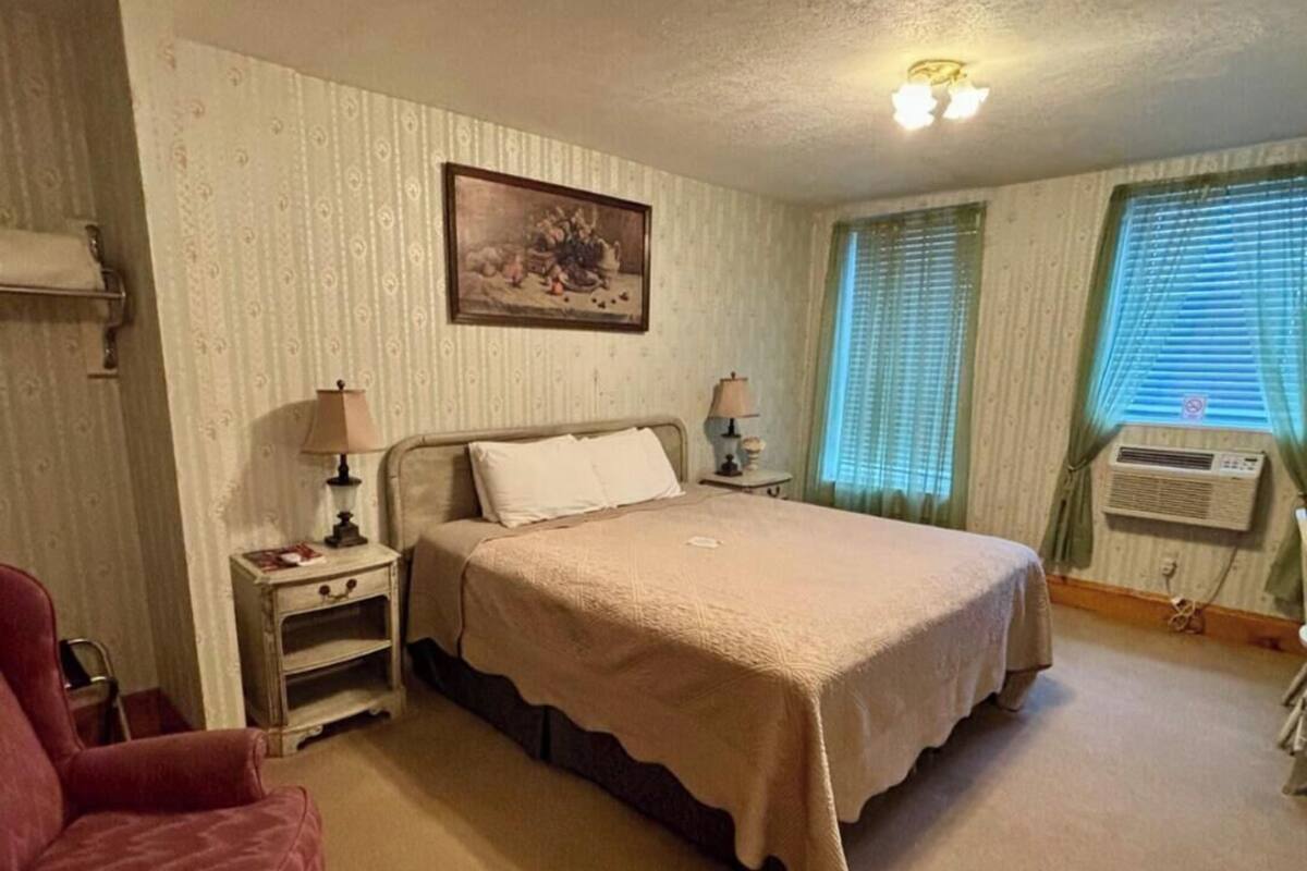 A bedroom with a bed, bedside tables, a lamp, a chair, a window with blinds, and an air conditioning unit.