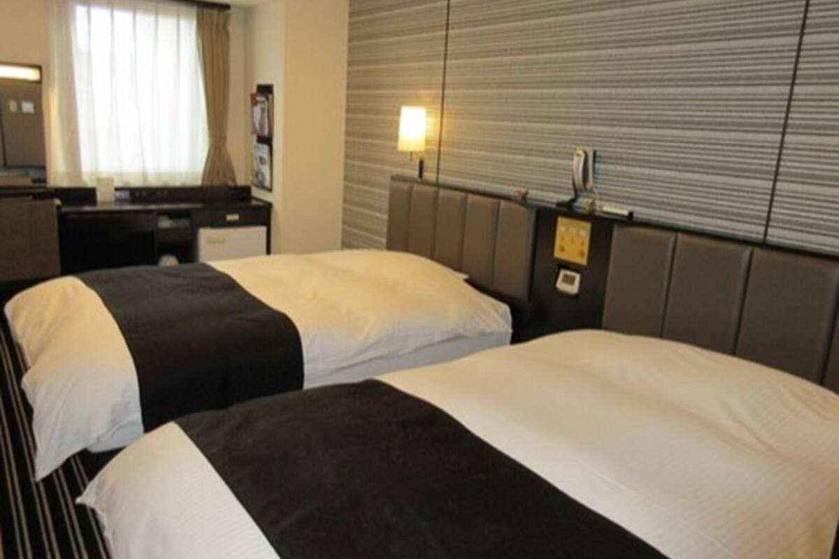 Habitación de hotel con dos camas, un escritorio, una lámpara y una ventana con cortinas.