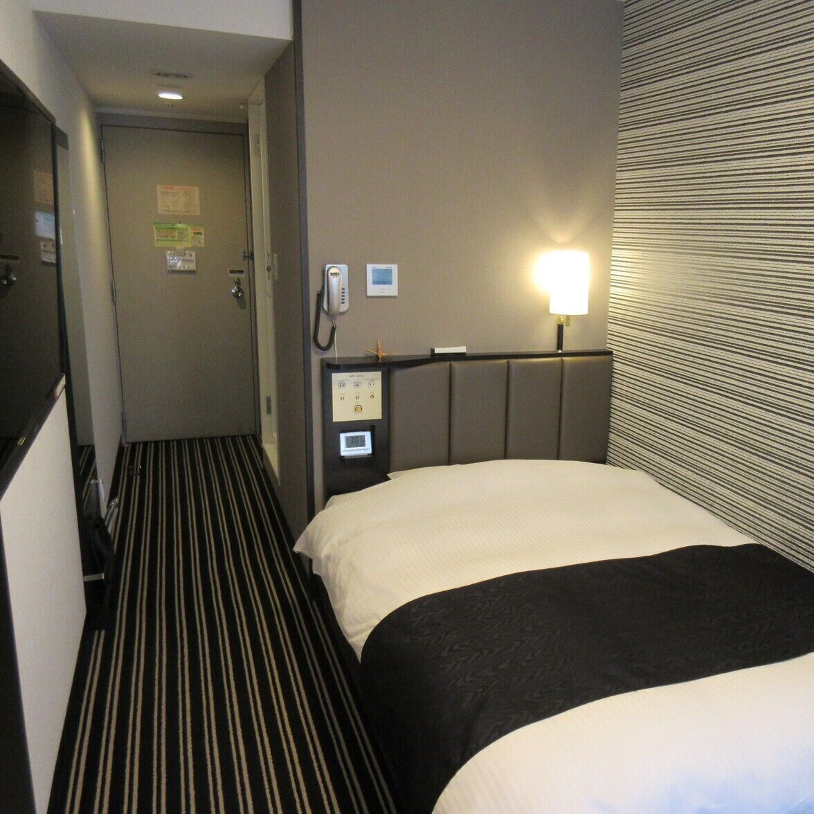 A hotel room with a bed, a bedside telephone, a control panel, and a door.