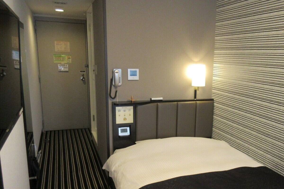 Una habitación de hotel con una cama, teléfono en la mesita de noche, panel de control y una puerta.