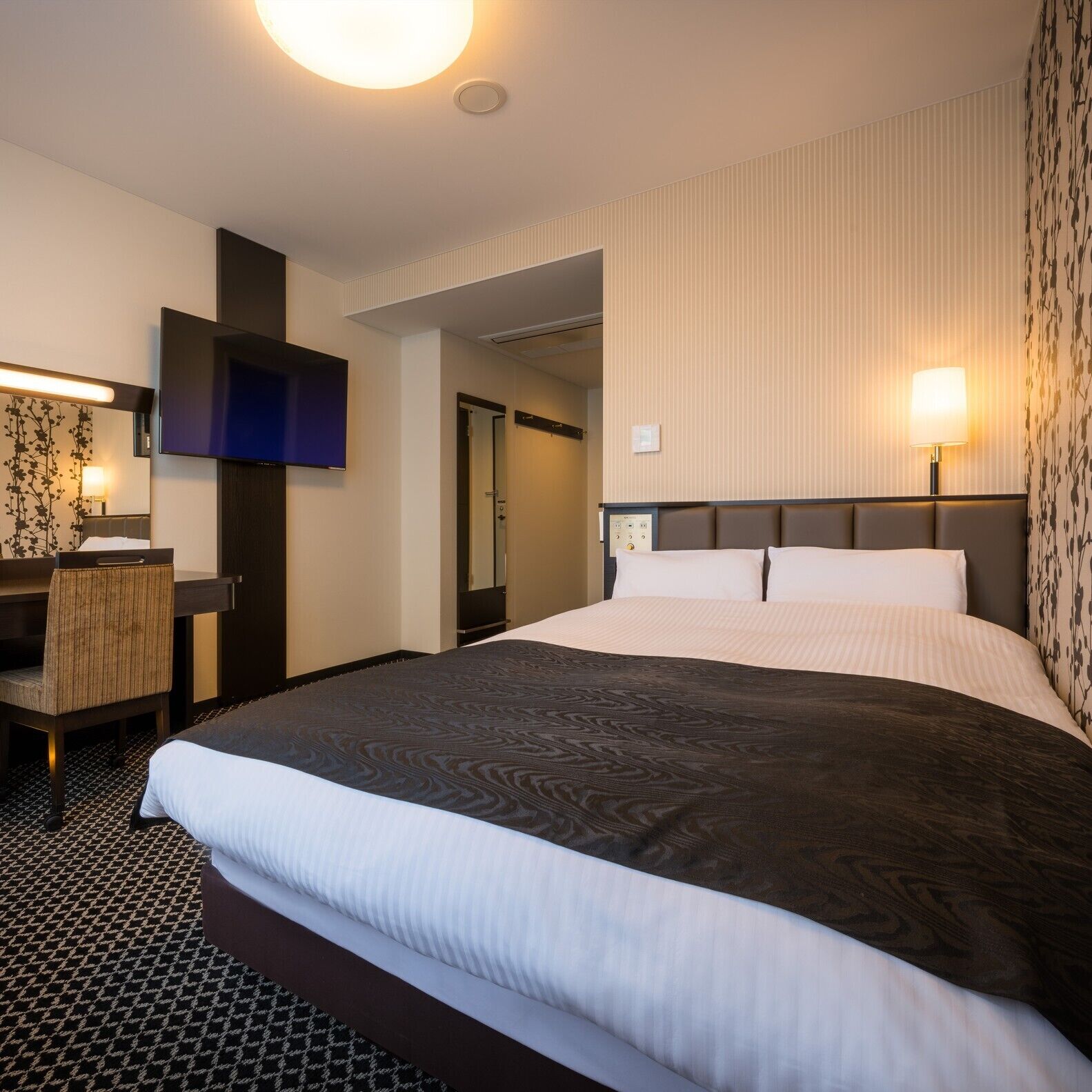 A hotel room with a large bed, a flat-screen TV, a desk with a chair, and a wall-mounted lamp.