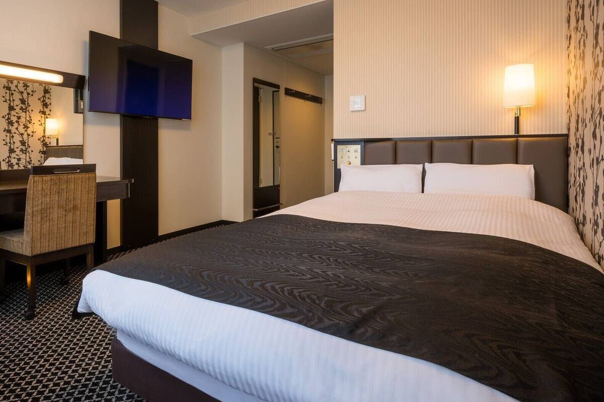 Una habitación de hotel con una cama grande, un televisor de pantalla plana, un escritorio con una silla y una lámpara de pared.