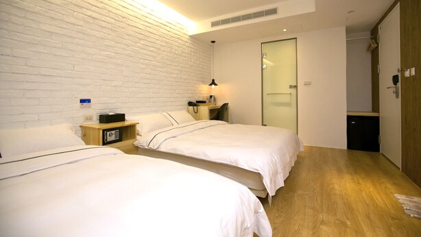 A hotel room with two beds, a wooden floor, a white brick wall, a desk with a lamp, and a door leading to a bathroom. - York Design Hotel (Taipei)