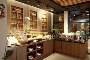 Café bem iluminado com prateleiras de madeira exibindo diversos objetos, balcão com louças e máquina de café, e uma grande janela com vista para o interior.