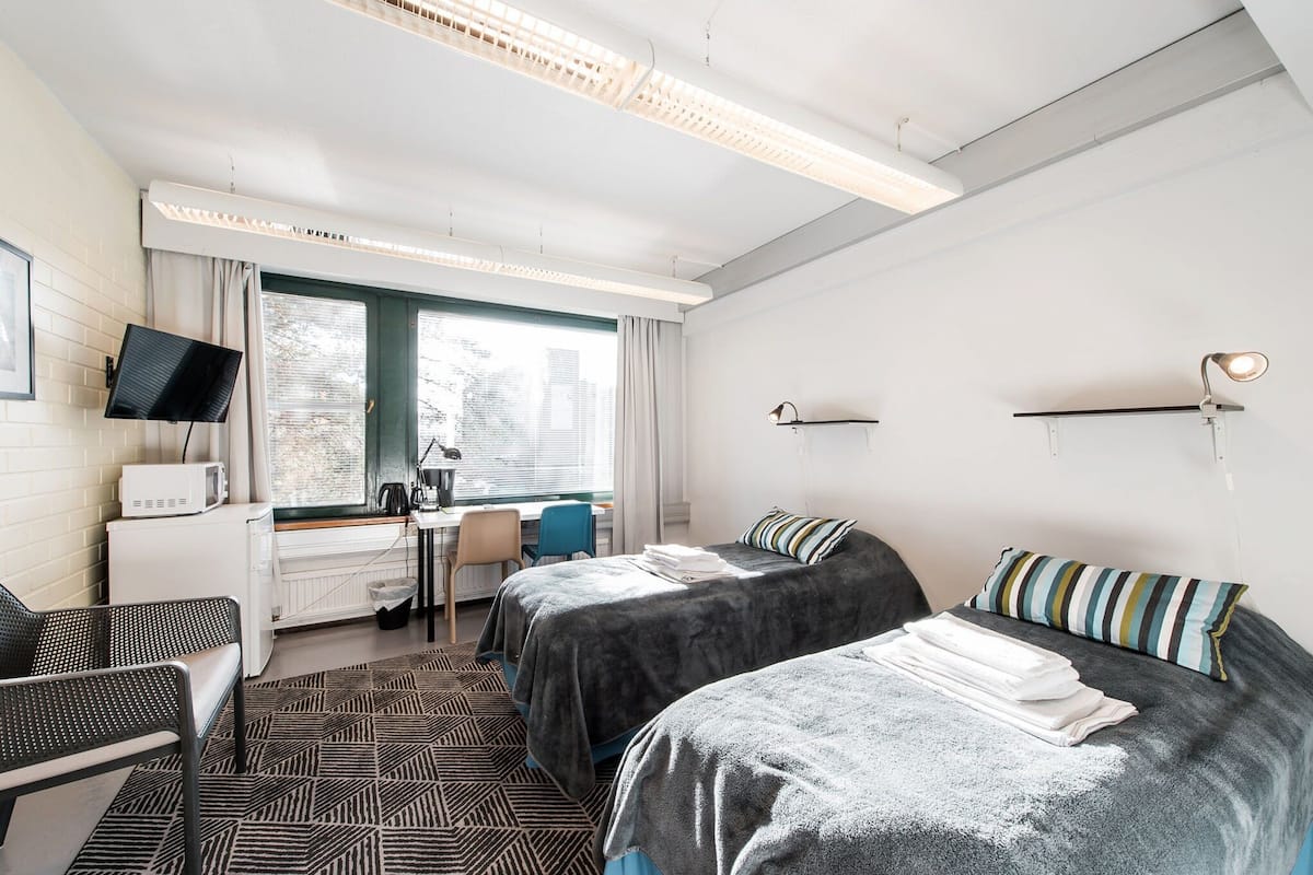 A hotel room with two beds, a desk, a chair, a TV, and a window with a view of buildings.
