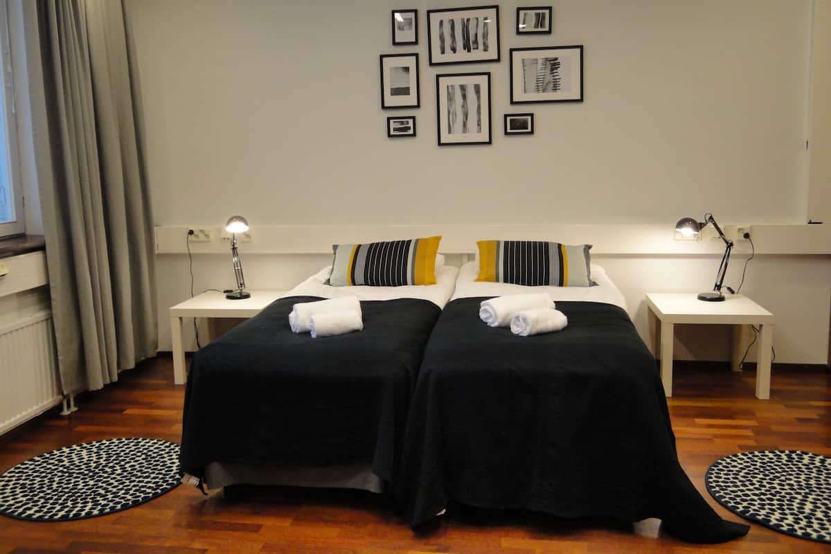 A hotel room with two beds, a nightstand with a lamp, a wall with framed pictures, and a window with curtains.