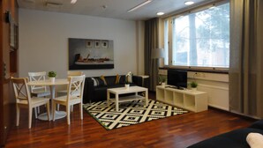 A modern living room with a wooden floor, a black sofa, a white coffee table, and a TV on a stand. There is a window with curtains, a painting on the wall, and a small table with a plant. - Forenom Hostel Espoo Otaniemi (Espoo)