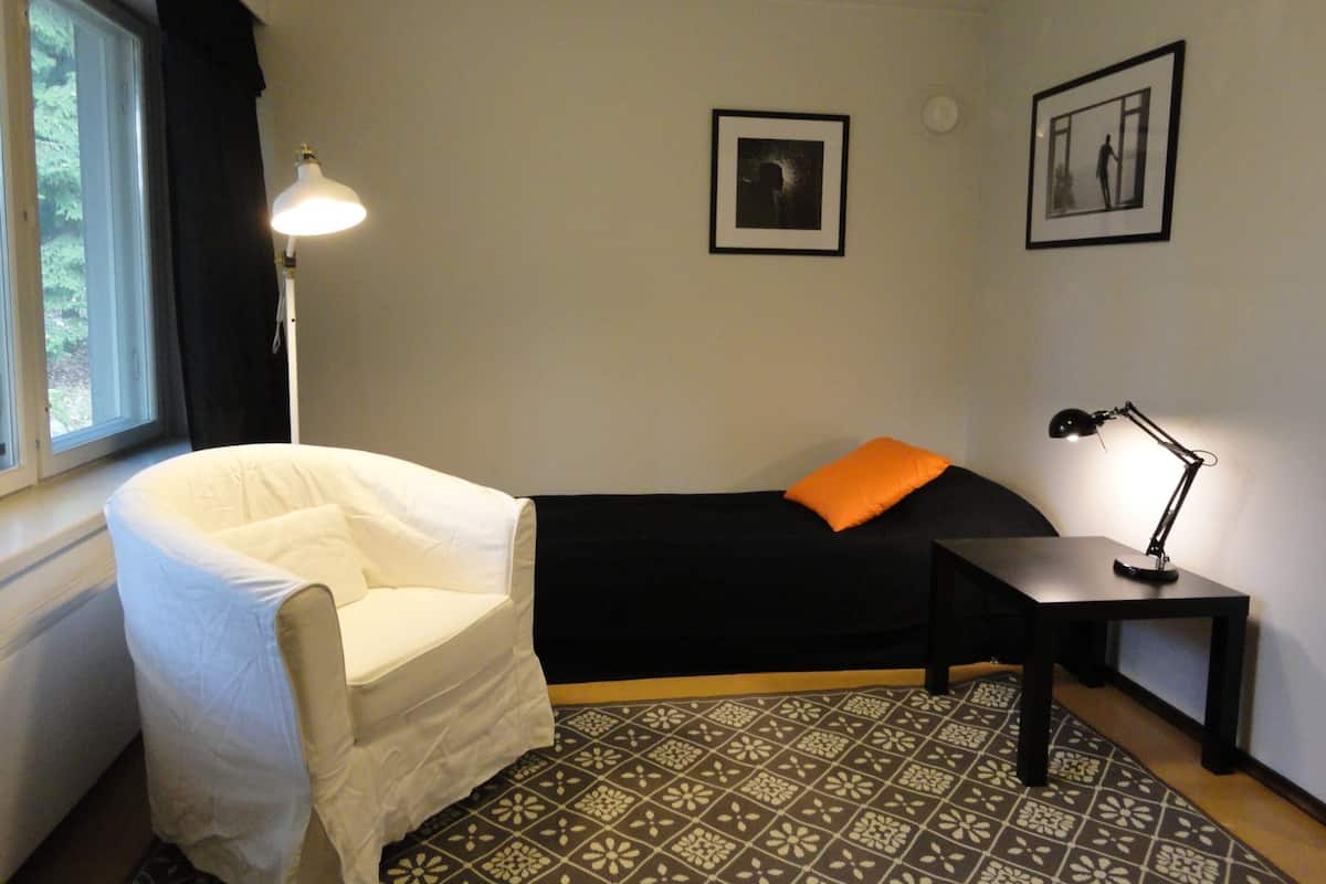 A small, neatly arranged bedroom with a single bed, a chair, a desk, and a lamp.