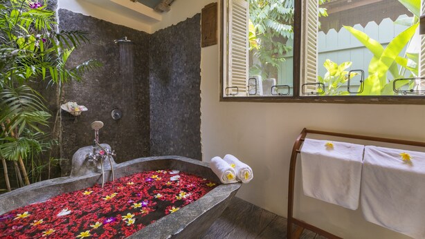 Ein Badezimmer mit einer steinernen Badewanne, gefüllt mit Wasser und Blütenblättern, einem Duschbereich und einem Fenster mit Blick auf Grünflächen.