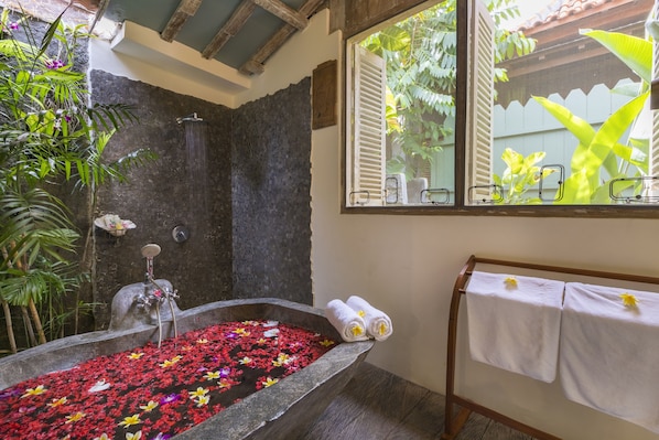 Un baño con una bañera de piedra llena de agua y pétalos de flores, una zona de ducha y una ventana con vistas a la vegetación.