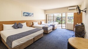 Habitación de hotel con tres camas, un escritorio, una silla y un televisor montado en la pared.