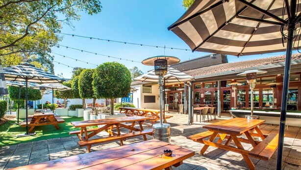 Un espacio de patio al aire libre con mesas de picnic de madera y sombrillas, rodeado de follaje y un edificio con techo de tejas.