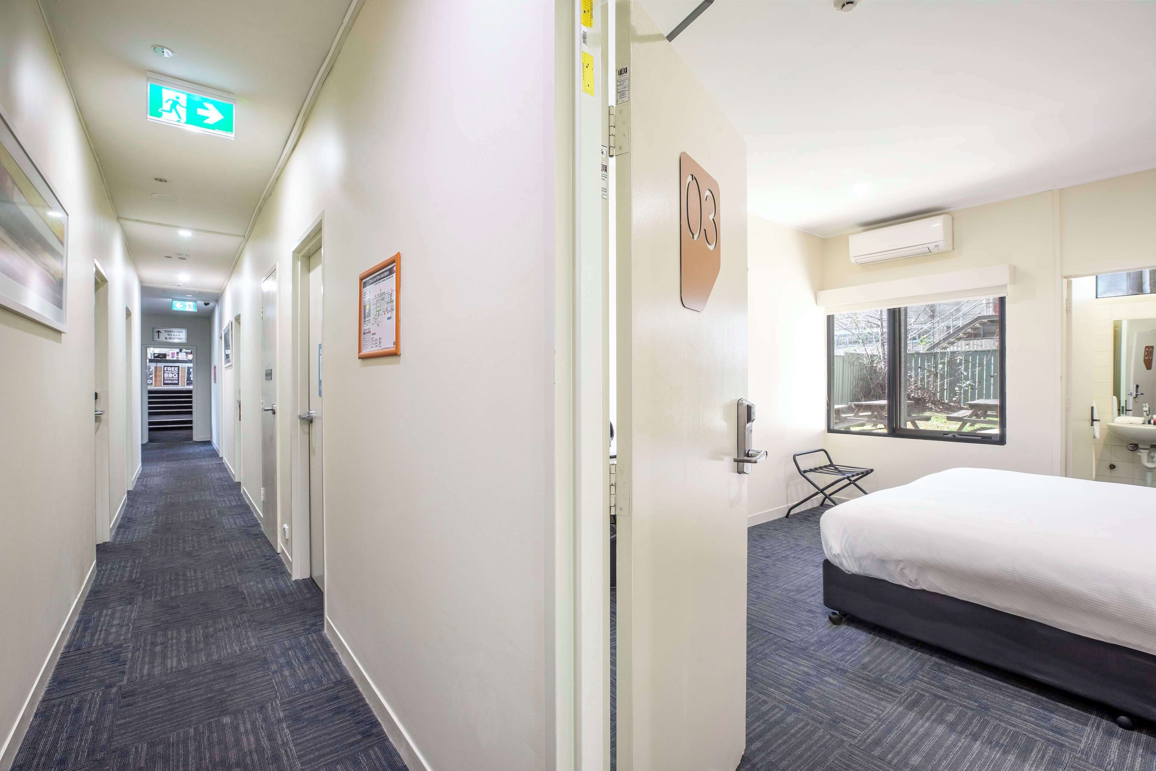 Een hotelgang met vloerbedekking, een bed met een witte matras, een raam met uitzicht en een vluchtroutebord.