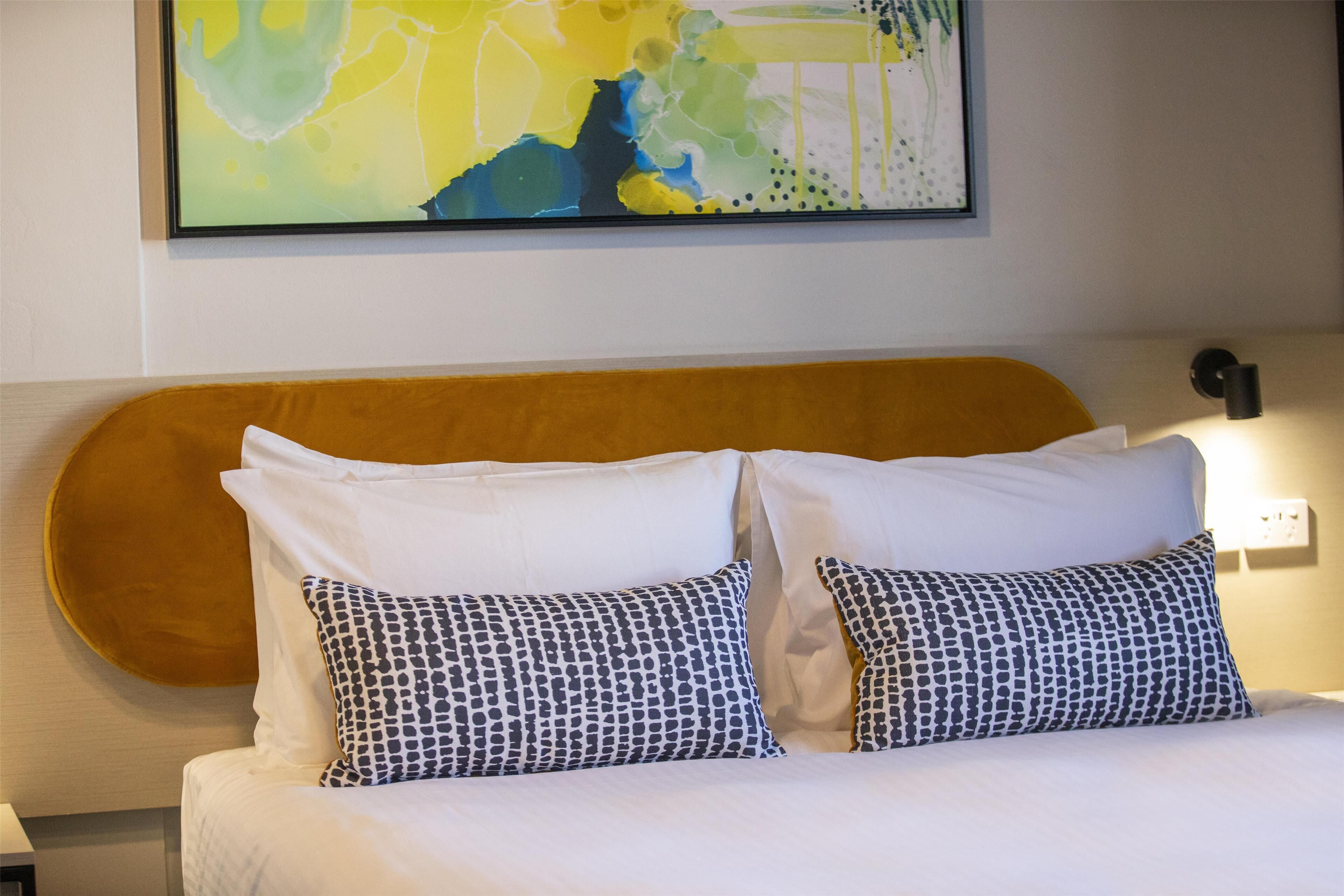 Una habitación de hotel con una cama, dos almohadas estampadas, un cabecero y un cuadro abstracto colgado en la pared.