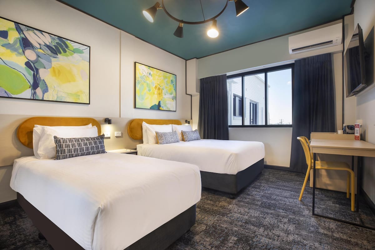 A hotel room with two beds, a desk, and a chair. There are two paintings on the walls, a window with curtains, and an air conditioning unit.