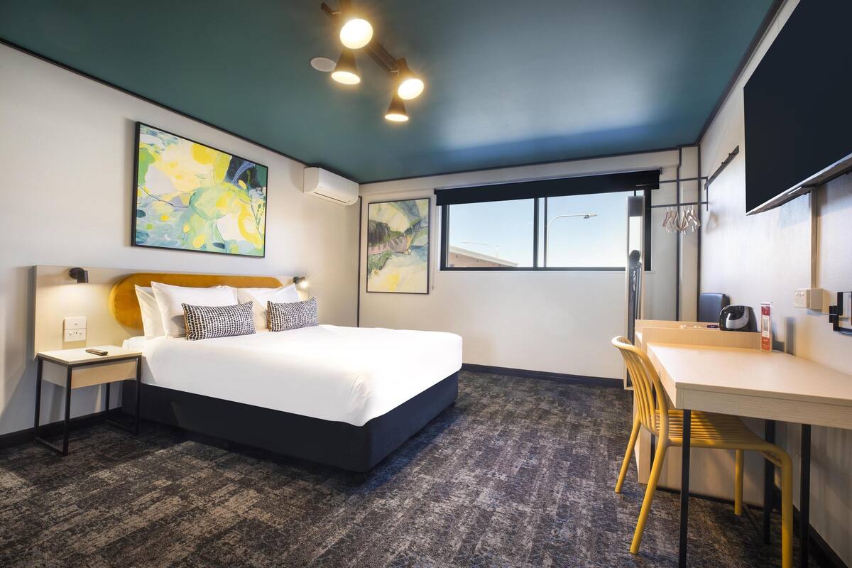 A hotel room with a bed, a desk, a chair, a window, and artwork on the walls.