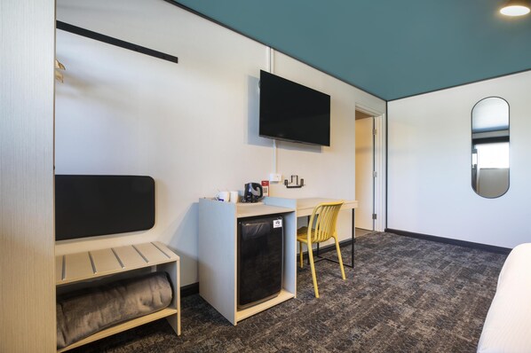 A compact hotel room with a bed, a desk, a small refrigerator, a flat-screen TV, and a mirror on the wall.