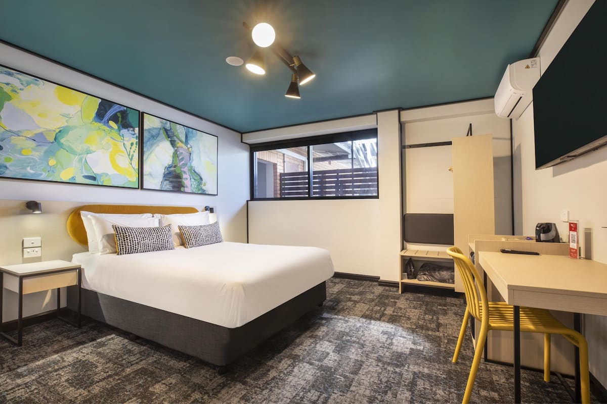 A modern hotel room with a bed, bedside table, and a desk with a chair. There are abstract paintings on the wall, a window with a view, and an air conditioning unit.