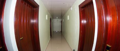 Un pasillo con dos puertas de madera, cada una con un número. El pasillo tiene piso de baldosas y un techo blanco con iluminación empotrada.