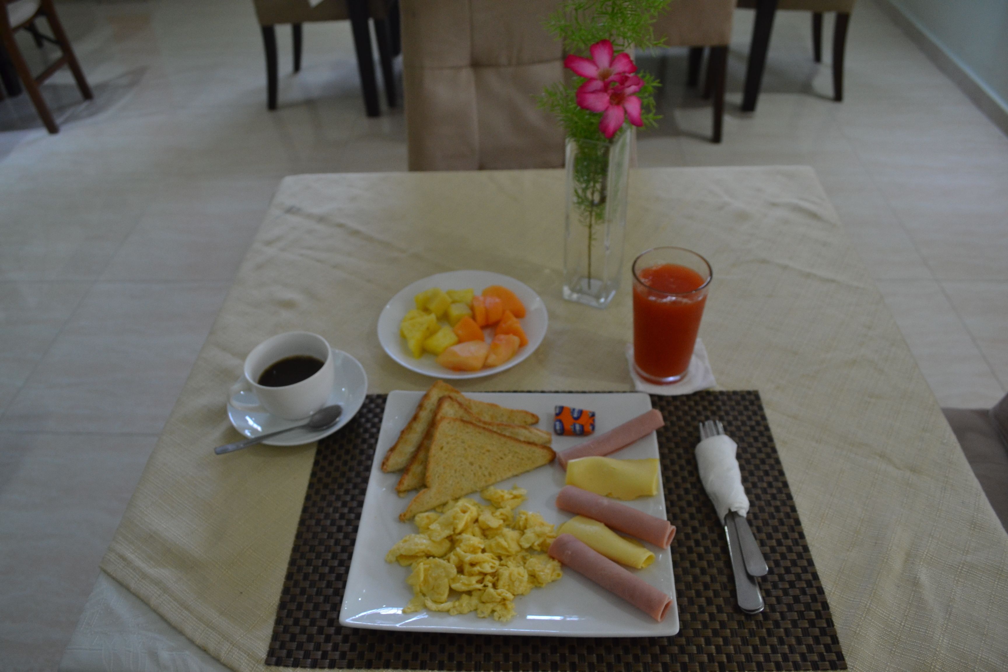 A breakfast table with toast, scrambled eggs, ham, and a plate of fruit. There is a glass of orange juice and a cup of coffee.