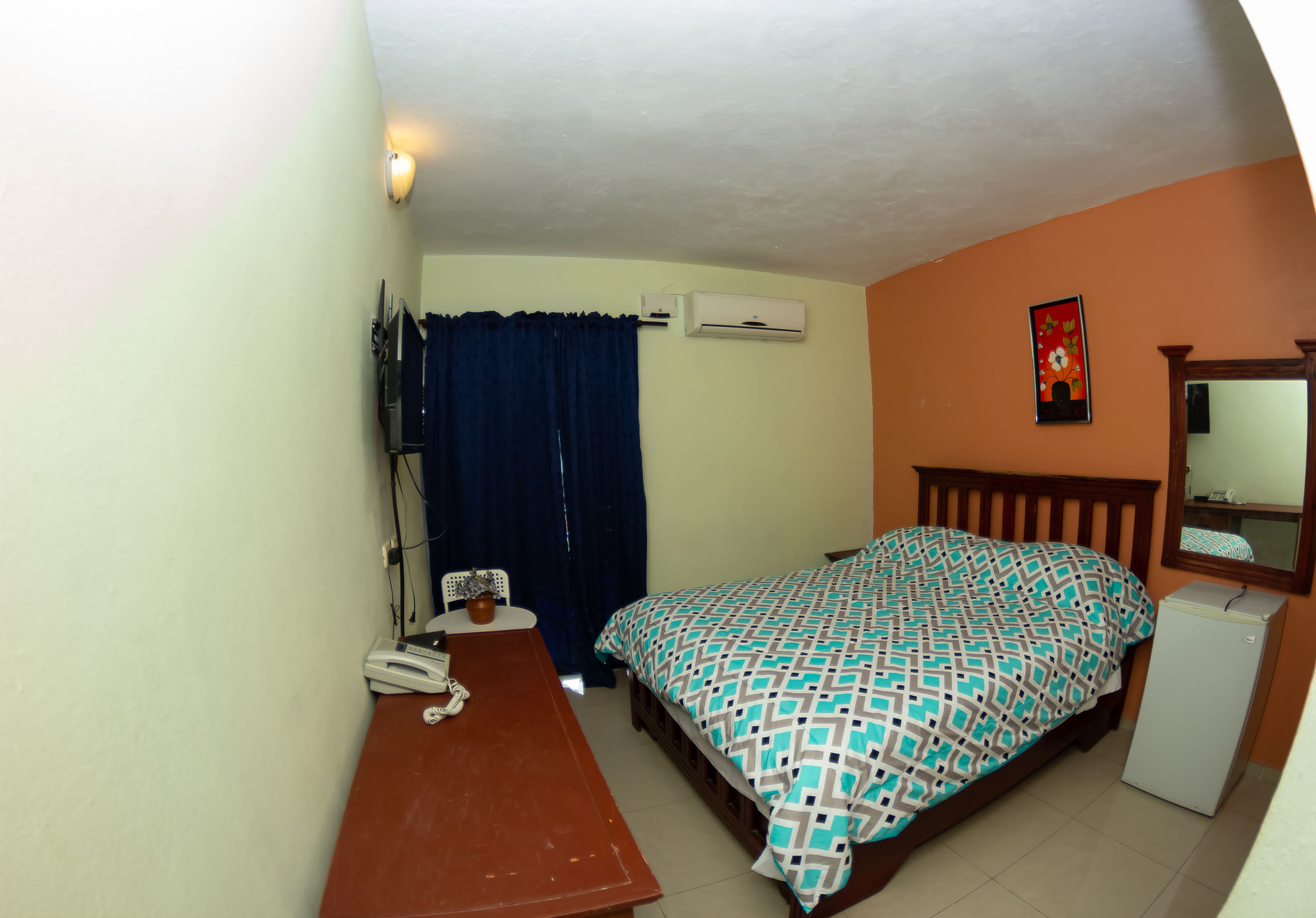 A bedroom with a bed, a desk, a chair, an air conditioner, a small refrigerator, and a window with curtains.