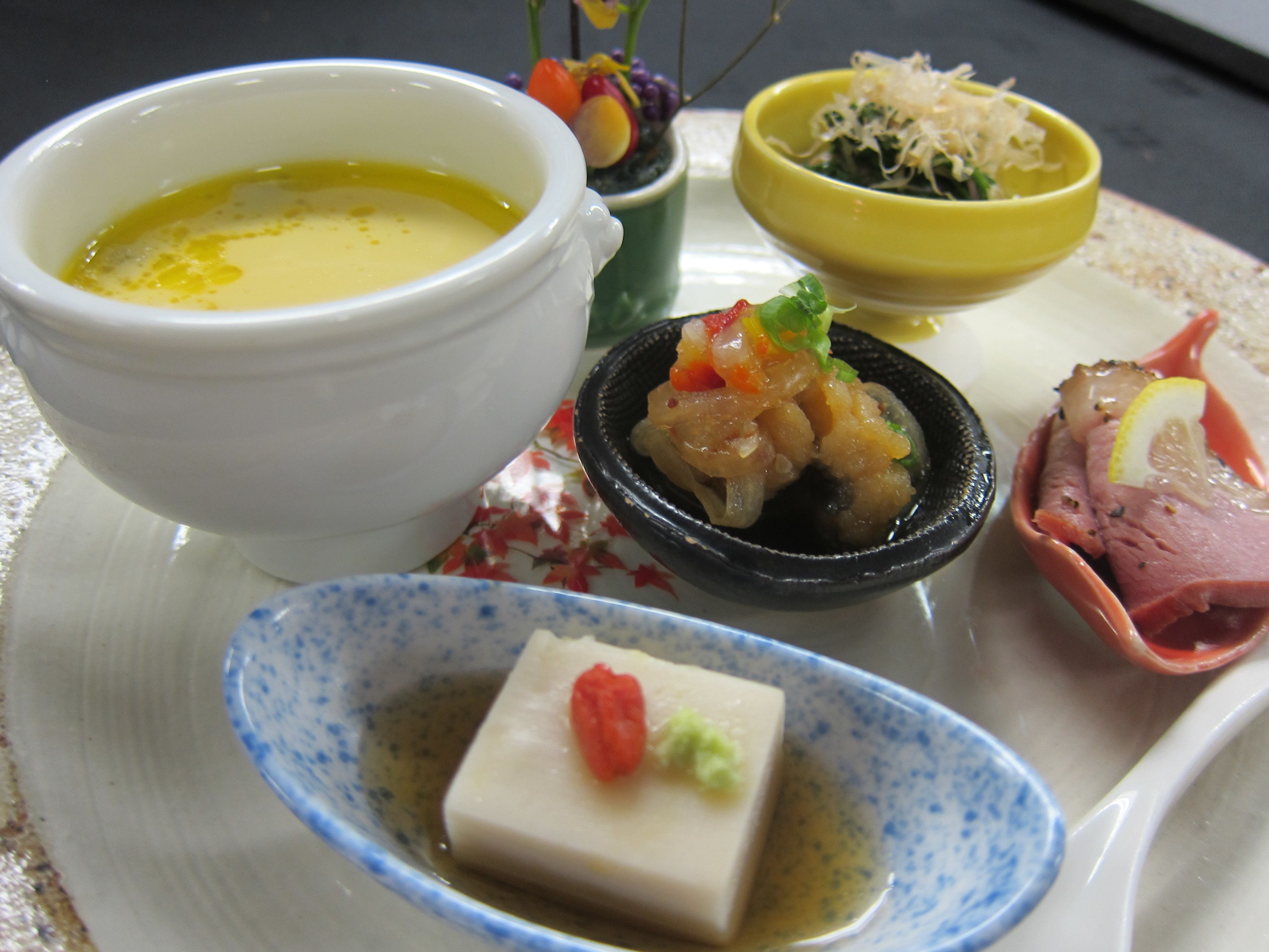 Variedad de platos, incluyendo un tazón de sopa, un recipiente pequeño de vegetales encurtidos, un plato con carne y vegetales, y un pequeño recipiente con tofu.