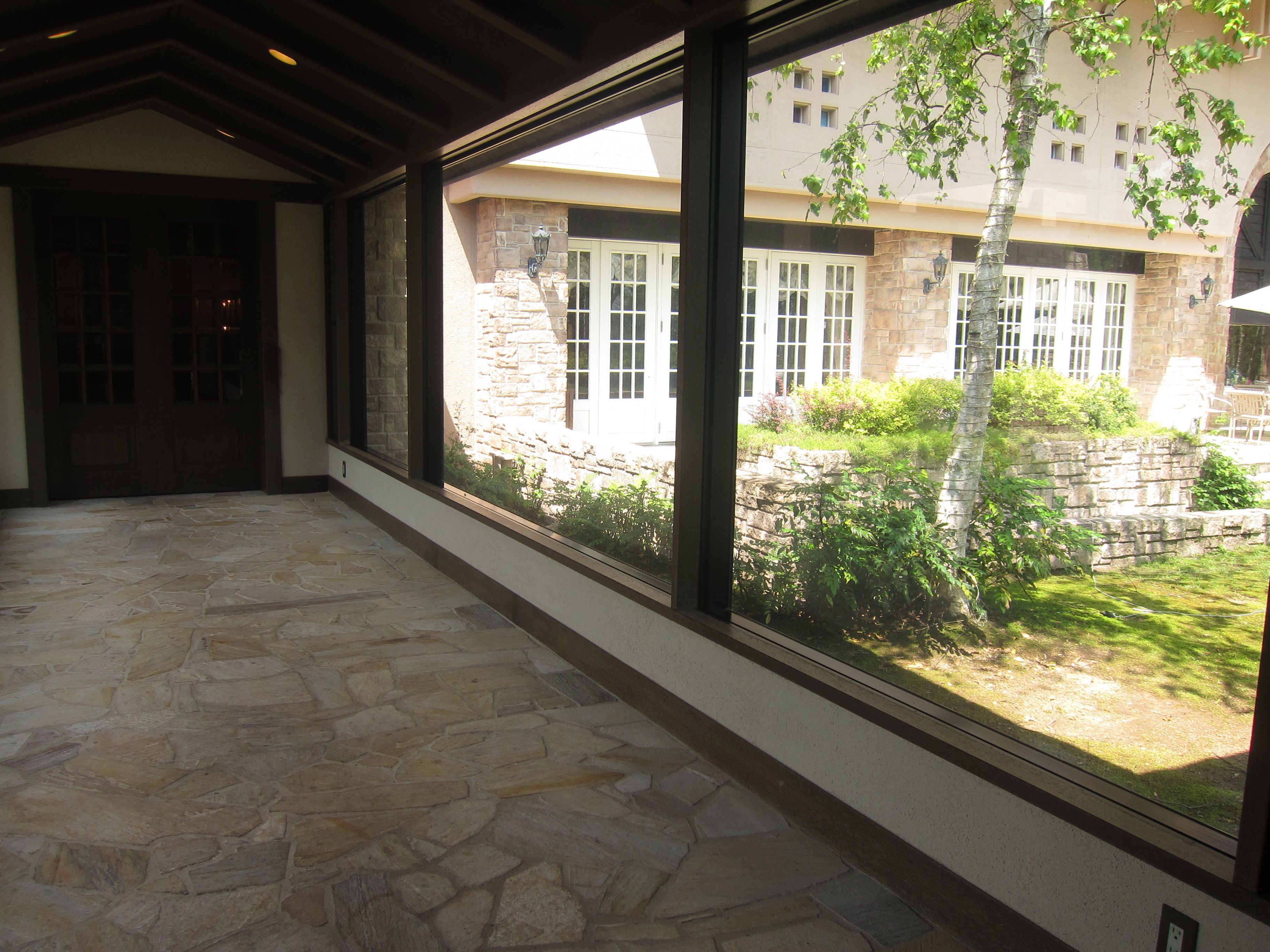 A hallway with stone flooring and large windows overlooking a courtyard with a stone wall and greenery.