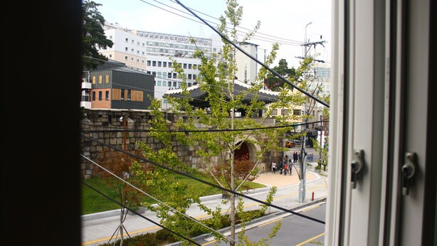A view through a window of a street with a stone wall, a tree, and buildings in the background. - Plan A Hostel (Seoul)