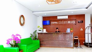 Un espace d’accueil d’hôtel avec un comptoir de réception en bois, une horloge numérique, un téléviseur et un canapé vert.