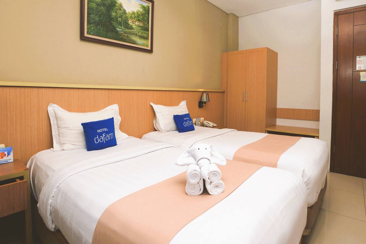 A hotel room with two beds, a wooden wardrobe, a painting on the wall, and a teddy bear on the bed.