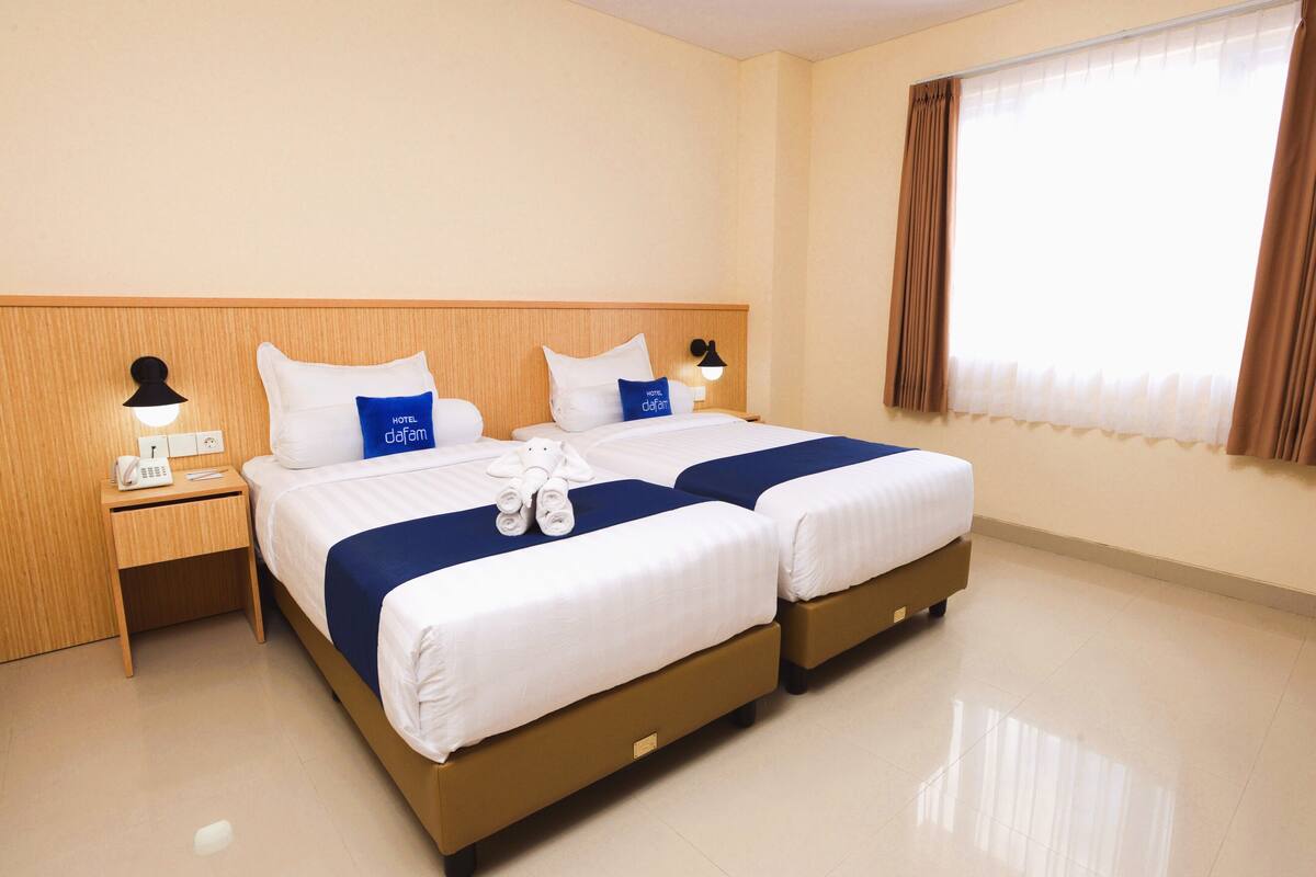 A hotel room with two beds, a wooden headboard, a nightstand with a phone and lamp, and a window with curtains.