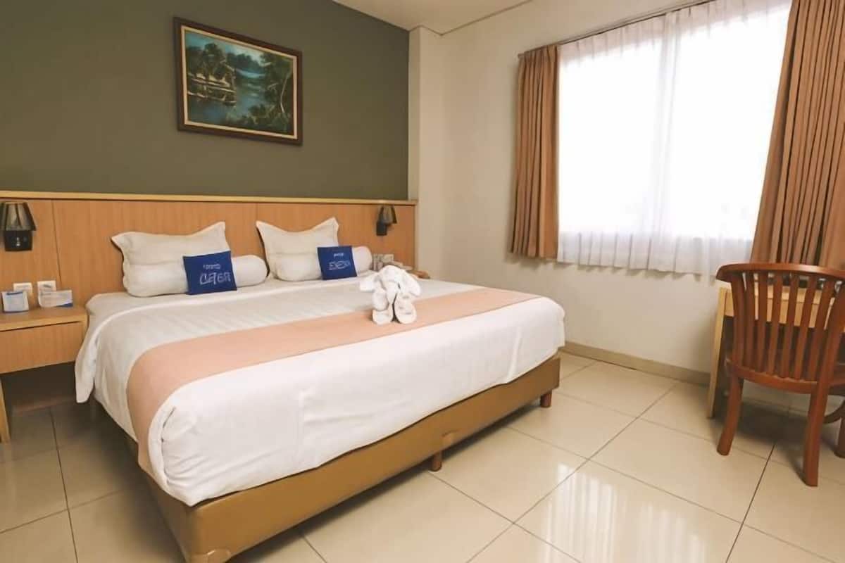 A hotel room with a bed, a wooden headboard, a nightstand, a chair, a window with curtains, and a painting on the wall.